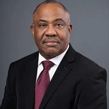 Professional portrait of a middle-aged man in a black suit, white shirt, and maroon tie against a gray background.