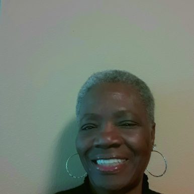A close-up portrait of a smiling elderly woman with short gray hair and hoop earrings against a plain background.