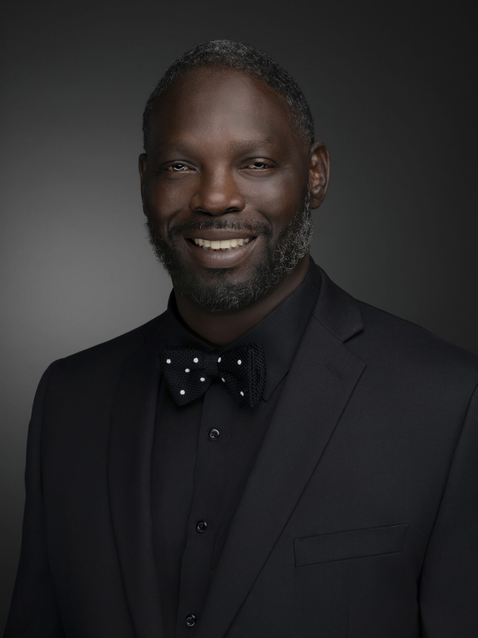 A smiling man wearing a black suit and a polka dot bow tie against a dark background.
